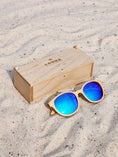 Load image into Gallery viewer, Maple Wooden sunglasses with blue polarized lenses and a box on sandy beach.