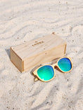 Load image into Gallery viewer, Maple Wooden sunglasses with green polarized lenses and a box on sandy beach.