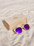 Load image into Gallery viewer, Maple Wooden sunglasses with purple polarized lenses and a box on sandy beach.