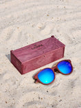 Load image into Gallery viewer, Purpleheart wooden sunglasses with blue polarized lenses and a box on sandy beach.