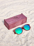 Load image into Gallery viewer, Purpleheart wooden sunglasses with green polarized lenses and a box on sandy beach.