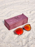 Load image into Gallery viewer, Purpleheart wooden sunglasses with red polarized lenses and a box on sandy beach.