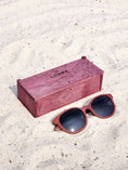 Load image into Gallery viewer, Purpleheart wooden sunglasses with black polarized lenses and a box on sandy beach.