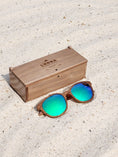 Load image into Gallery viewer, Walnut wooden sunglasses with green polarized lenses and a box on sandy beach.