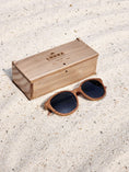 Load image into Gallery viewer, Walnut wooden sunglasses with black polarized lenses and a box on sandy beach.