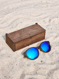 Load image into Gallery viewer, Wenge wooden sunglasses with blue polarized lenses and a box on sandy beach.