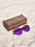 Load image into Gallery viewer, Wenge wooden sunglasses with purple polarized lenses and a box on sandy beach.