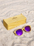 Load image into Gallery viewer, Yellowheart wooden sunglasses with purple polarized lenses and a box on sandy beach.