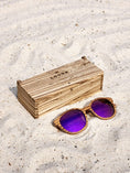 Load image into Gallery viewer, Zebrawood wooden sunglasses with purple polarized lenses and a box on sandy beach.