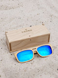 Load image into Gallery viewer, Maple wooden sunglasses with blue polarized lenses and a box on sandy beach.