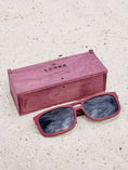 Load image into Gallery viewer, Purpleheart wooden sunglasses with black polarized lenses and a box on sandy beach.