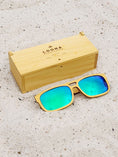 Load image into Gallery viewer, Yellowheart wooden sunglasses with green polarized lenses and a box on sandy beach.