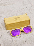 Load image into Gallery viewer, Yellowheart wooden sunglasses with purple polarized lenses and a box on sandy beach.