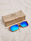 Load image into Gallery viewer, Zebrawood wooden sunglasses with blue polarized lenses and a box on sandy beach.