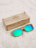 Load image into Gallery viewer, Zebrawood wooden sunglasses with green polarized lenses and a box on sandy beach.