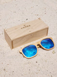 Load image into Gallery viewer, Maple wooden sunglasses with blue polarized lenses and a box on sandy beach.