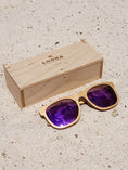 Load image into Gallery viewer, Maple wooden sunglasses with purple polarized lenses and a box on sandy beach.
