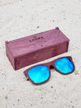 Load image into Gallery viewer, Purpleheart wooden sunglasses with blue polarized lenses and a box on sandy beach.