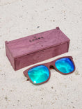 Load image into Gallery viewer, Purpleheart wooden sunglasses with green polarized lenses and a box on sandy beach.