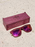 Load image into Gallery viewer, Purpleheart wooden sunglasses with red polarized lenses and a box on sandy beach.
