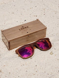 Load image into Gallery viewer, Walnut wooden sunglasses with red polarized lenses and a box on sandy beach.