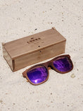 Load image into Gallery viewer, Walnut wooden sunglasses with purple polarized lenses and a box on sandy beach.