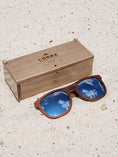 Load image into Gallery viewer, Walnut wooden sunglasses with silver polarized lenses and a box on sandy beach.