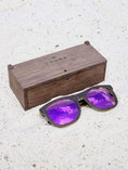 Load image into Gallery viewer, Wenge wooden sunglasses with purple polarized lenses and a box on sandy beach.