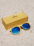 Load image into Gallery viewer, Yellowheart wooden sunglasses with blue polarized lenses and a box on sandy beach.