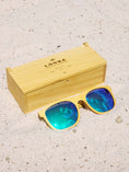 Load image into Gallery viewer, Yellowheart wooden sunglasses with green polarized lenses and a box on sandy beach.