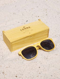 Load image into Gallery viewer, Yellowheart wooden sunglasses with black polarized lenses and a box on sandy beach.