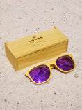 Load image into Gallery viewer, Yellowheart wooden sunglasses with purple polarized lenses and a box on sandy beach.