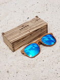 Load image into Gallery viewer, Zebrawood wooden sunglasses with blue polarized lenses and a box on sandy beach.