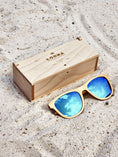 Load image into Gallery viewer, Maple wooden sunglasses with blue polarized lenses and a box on sandy beach.