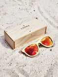 Load image into Gallery viewer, Maple wooden sunglasses with red polarized lenses and a box on sandy beach.