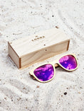 Load image into Gallery viewer, Maple wooden sunglasses with purple polarized lenses and a box on sandy beach.