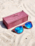 Load image into Gallery viewer, Purpleheart wooden sunglasses with blue polarized lenses and a box on sandy beach.