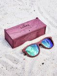 Load image into Gallery viewer, Purpleheart wooden sunglasses with green polarized lenses and a box on sandy beach.