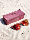 Load image into Gallery viewer, Purpleheart wooden sunglasses with red polarized lenses and a box on sandy beach.