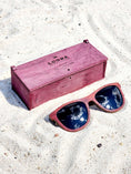 Load image into Gallery viewer, Purpleheart wooden sunglasses with black polarized lenses and a box on sandy beach.