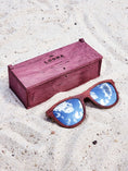 Load image into Gallery viewer, Purpleheart wooden sunglasses with silver polarized lenses and a box on sandy beach.