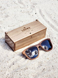 Load image into Gallery viewer, Walnut wooden sunglasses with black polarized lenses and a box on sandy beach.