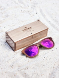 Load image into Gallery viewer, Walnut wooden sunglasses with purple polarized lenses and a box on sandy beach.