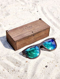 Load image into Gallery viewer, Wenge wooden sunglasses with green polarized lenses and a box on sandy beach.