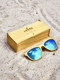 Load image into Gallery viewer, Yellowheart wooden sunglasses with blue polarized lenses and a box on sandy beach.