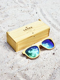 Load image into Gallery viewer, Yellowheart wooden sunglasses with green polarized lenses and a box on sandy beach.