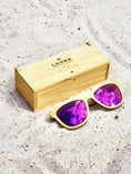 Load image into Gallery viewer, Yellowheart wooden sunglasses with purple polarized lenses and a box on sandy beach.