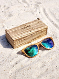 Load image into Gallery viewer, Zebrawood wooden sunglasses with green polarized lenses and a box on sandy beach.
