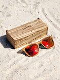 Load image into Gallery viewer, Zebrawood wooden sunglasses with red polarized lenses and a box on sandy beach.
