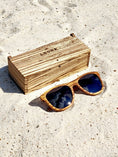 Load image into Gallery viewer, Zebrawood wooden sunglasses with black polarized lenses and a box on sandy beach.